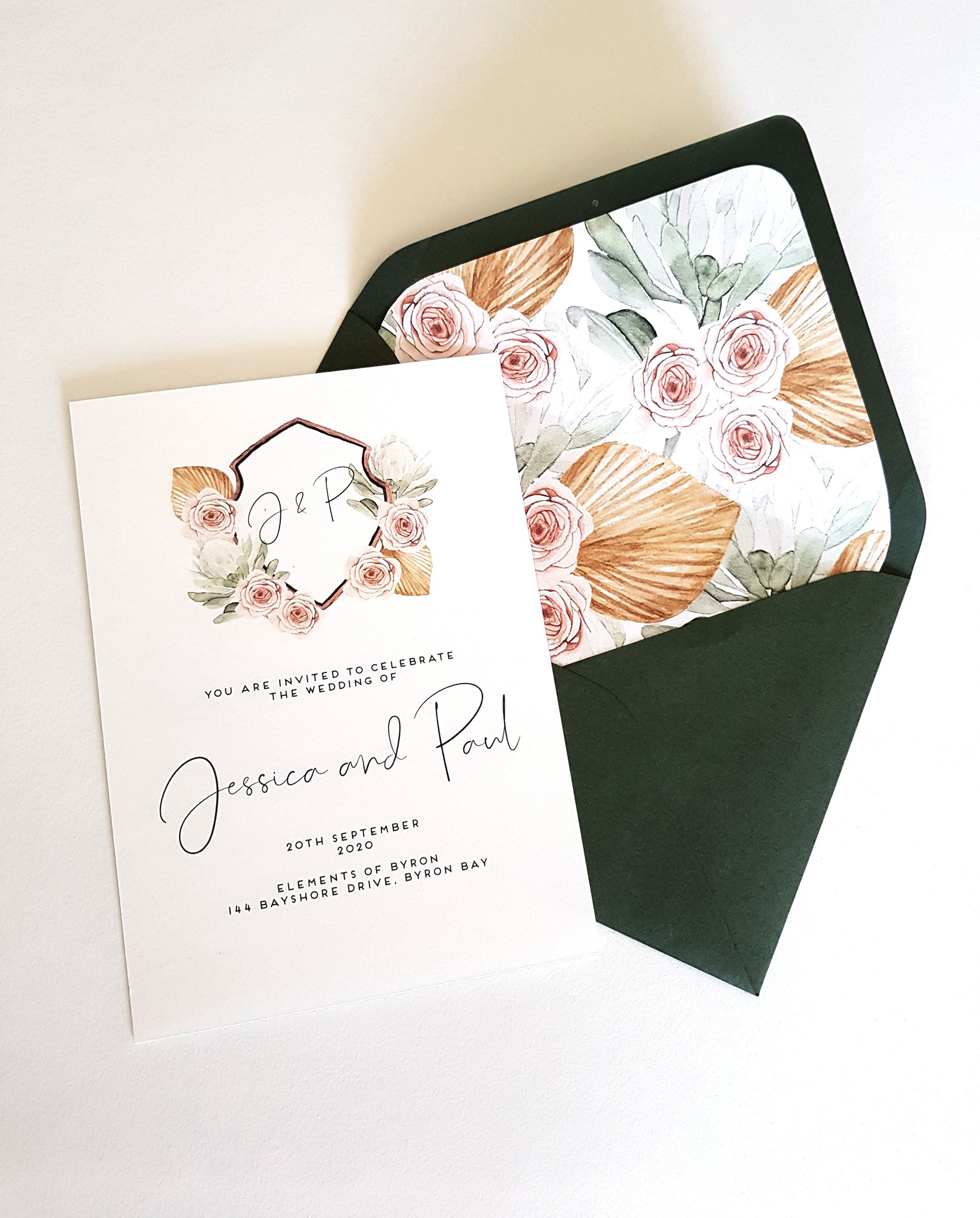 Crest wedding invite design, featuring wild roses in dusty pink, green protea, tan coloured tropical leaves, around a hand painted crest and green envelope